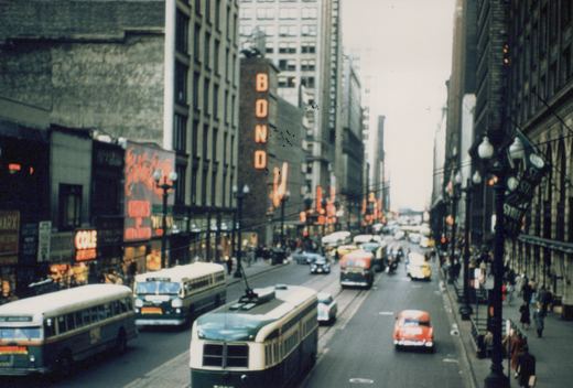 Elevated view along a busy downtown street lined with neon signs: a green and cream streetcar and two green and cream buses are in the foreground, with cars, a taxi and a red-fronted bus behind them.