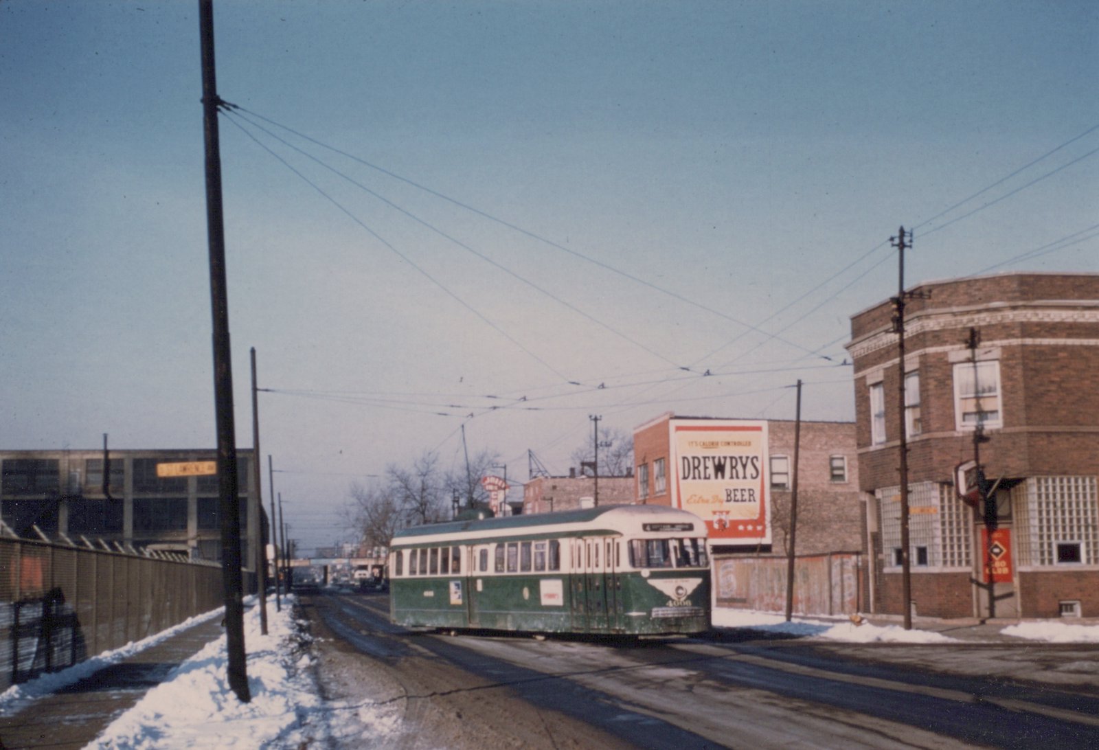 A green and cream streetcar rounds a snowy street corner under overhead wires, next to a brick tavern and a painted Drewrys Beer sign.