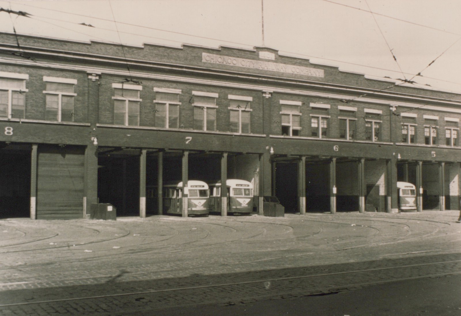 A two-story brick carbarn lettered Chicago City Railway Company with numbered open bays. Three streamlined streetcars are parked inside, with curving tracks and overhead wires in front.