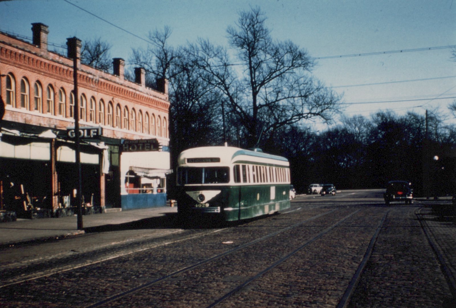 A green and cream streetcar numbered 4029 runs on a cobbled street with two sets of tracks, beside a two-story brick building with arched windows and GOLF and HOTEL signs, with bare trees and overhead wires behind it.