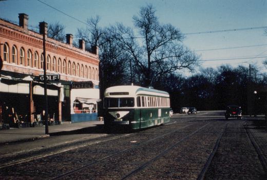 A green and cream streetcar numbered 4029 runs on a cobbled street with two sets of tracks, beside a two-story brick building with arched windows and GOLF and HOTEL signs, with bare trees and overhead wires behind it.