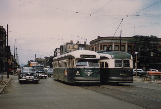 Two green and cream streetcars with trolley poles stand side by side on a street under overhead wires, with 1950s cars and trucks at the left and brick buildings behind.
