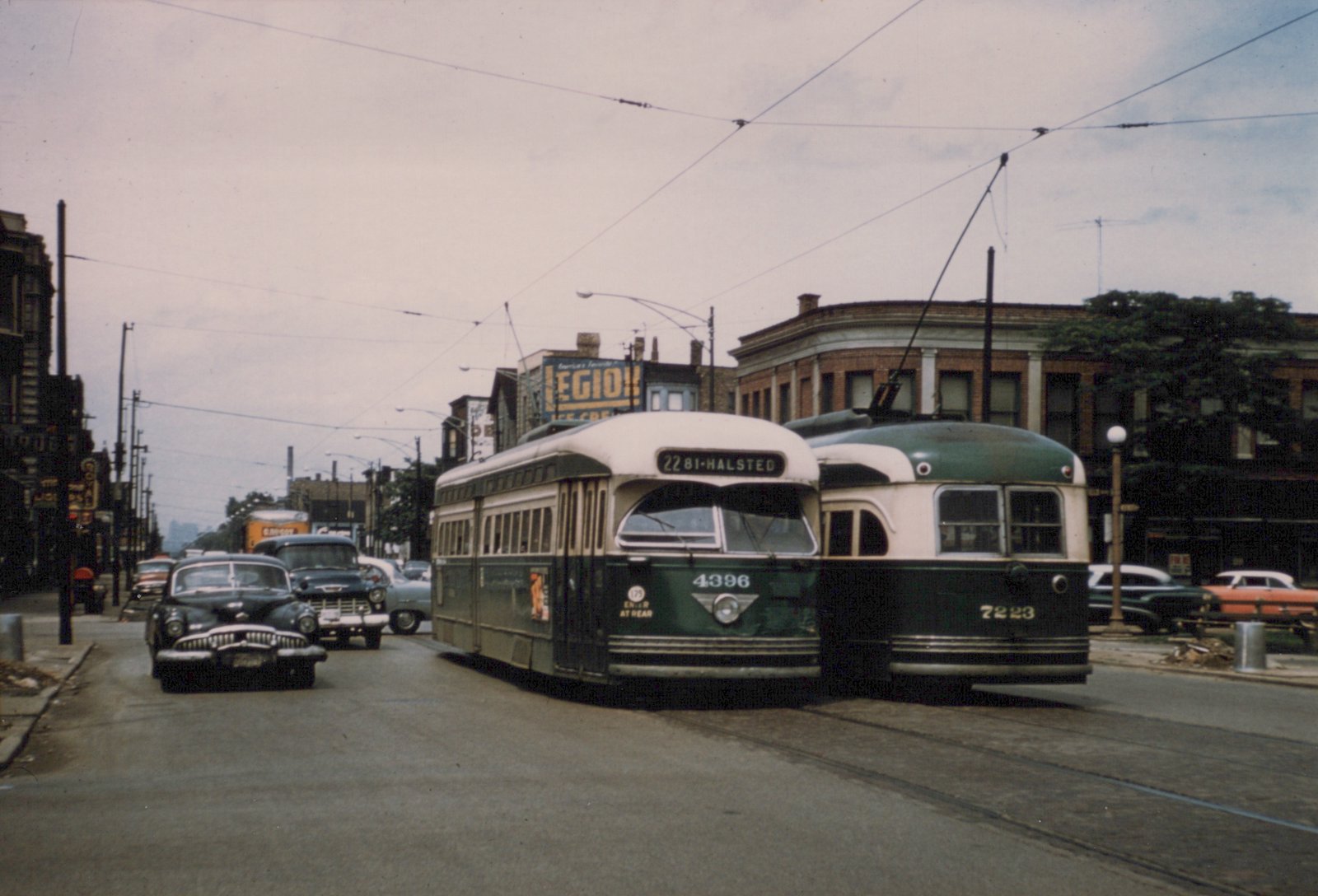 Two green and cream streetcars with trolley poles stand side by side on a street under overhead wires, with 1950s cars and trucks at the left and brick buildings behind.