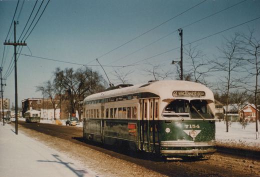 A green and cream streetcar with snow on its roof travels toward the camera on a slushy street lined with bare trees and snow-covered lawns, with another streetcar farther back.