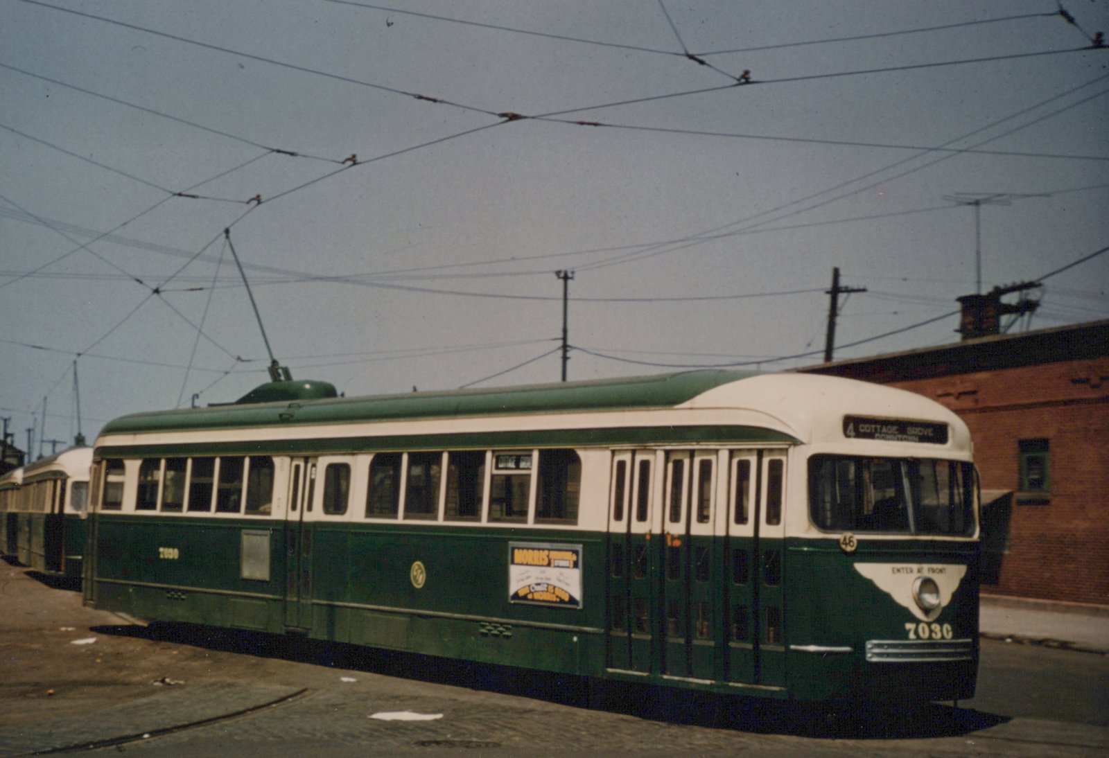 A green and cream streetcar numbered 7030 with a trolley pole up stands on brick-paved track under a web of overhead wires, with another streetcar behind it and a red brick building at right.