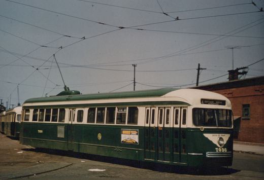 A green and cream streetcar numbered 7030 with a trolley pole up stands on brick-paved track under a web of overhead wires, with another streetcar behind it and a red brick building at right.