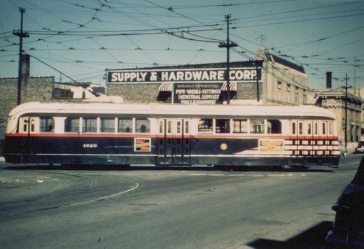 Side view of a dark green and cream streamlined streetcar with a red stripe, numbered 4020, on curving track in front of a brick building with a large painted sign reading Supply & Hardware Corp., beneath a dense web of overhead wires.