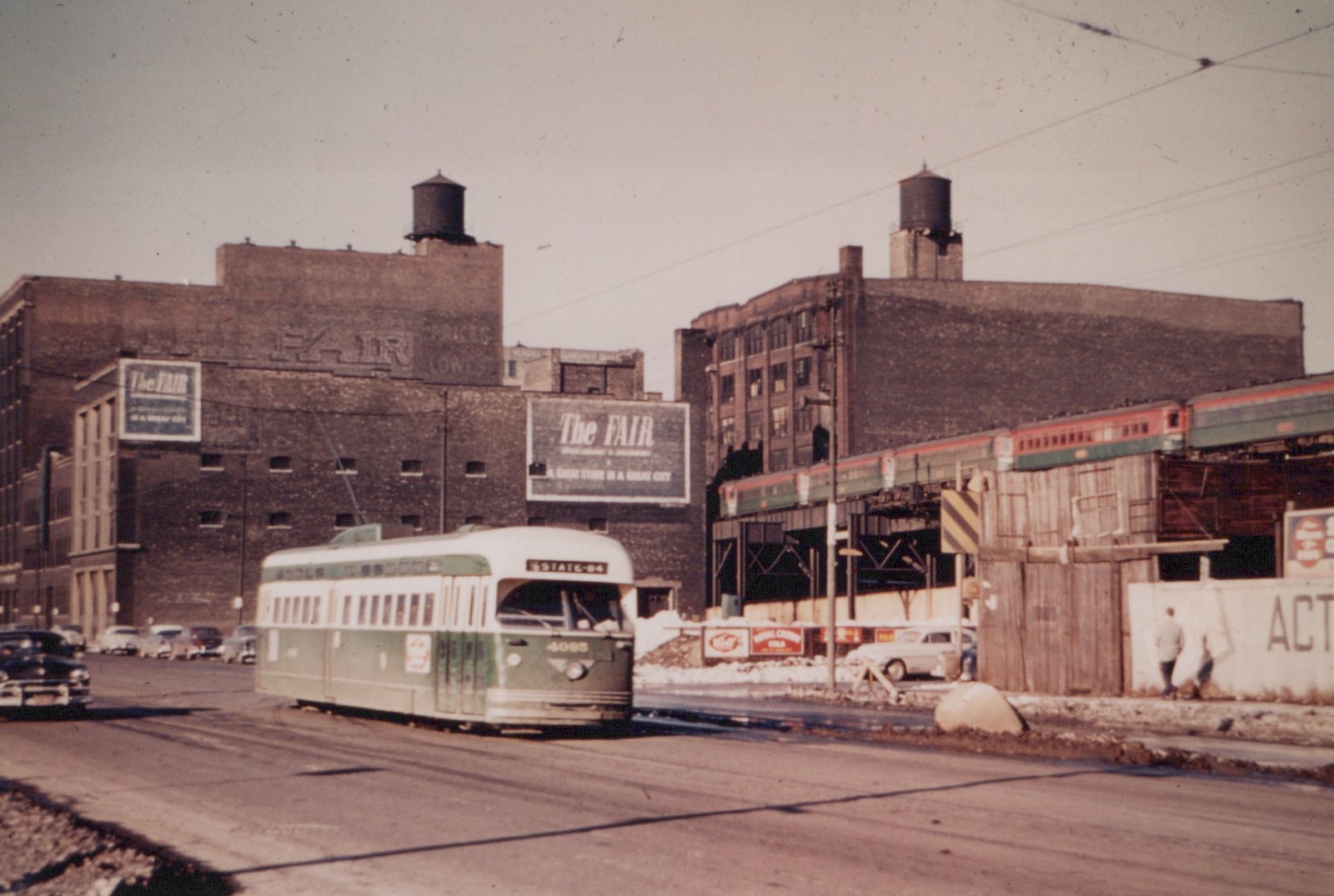 A green and cream streetcar numbered 4095 travels down a slushy city street past brick warehouses with The Fair billboards, while a red and green train passes on an elevated structure at right.
