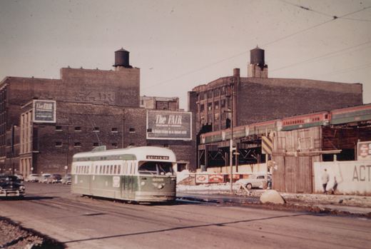 A green and cream streetcar numbered 4095 travels down a slushy city street past brick warehouses with The Fair billboards, while a red and green train passes on an elevated structure at right.