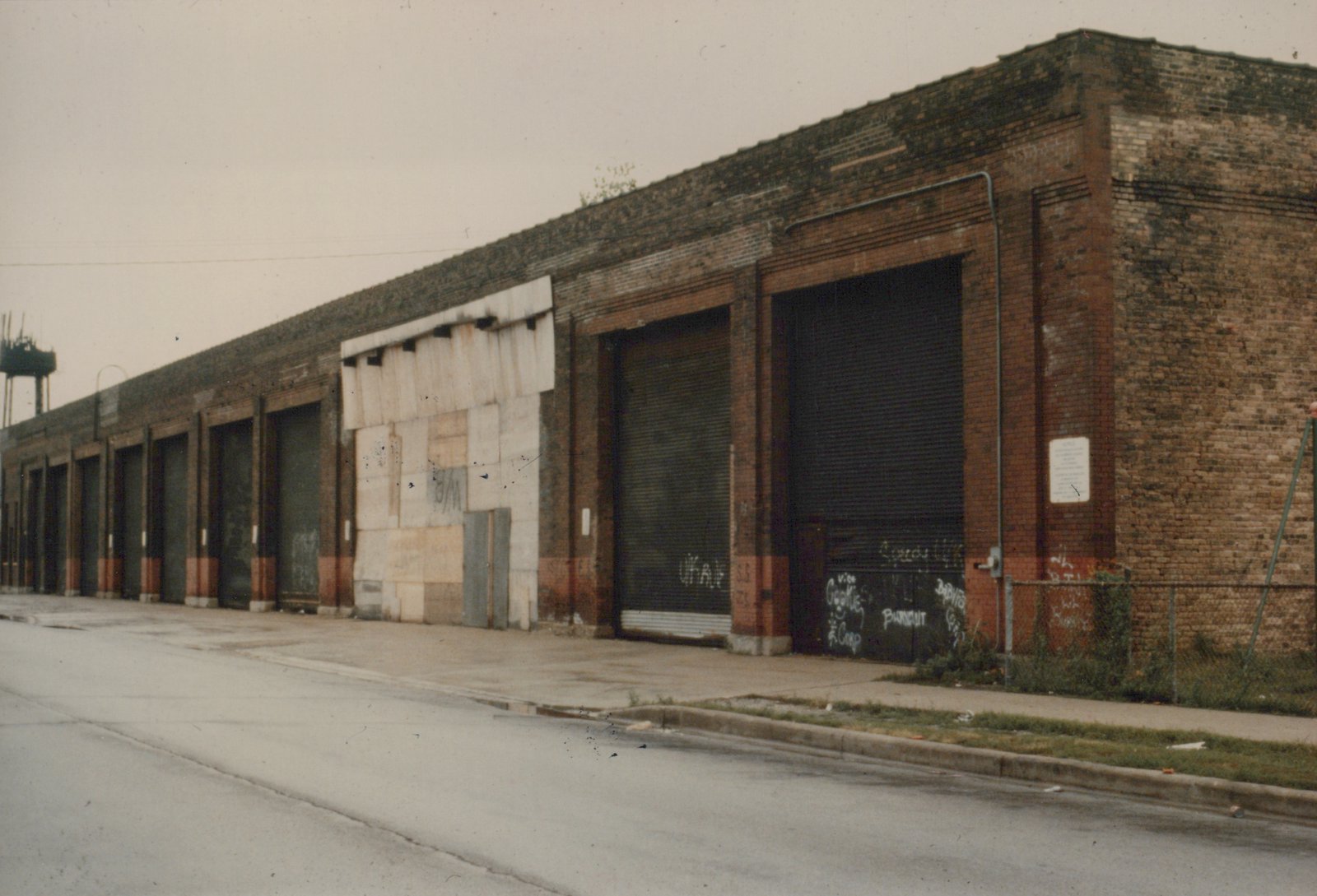 A long, one-story brown brick building with many closed roll-up doors and one boarded-up bay, graffiti on the doors, a wet street in the foreground and an overcast sky.