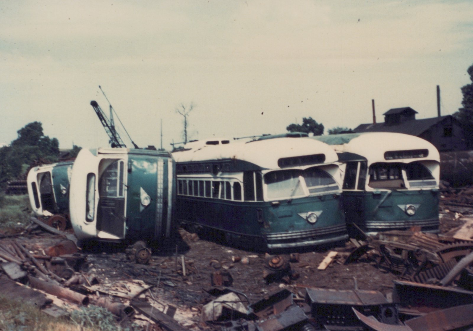 Four green and cream streamlined streetcars without their trucks, two lying on their sides and two upright, in a yard full of scrap metal, with a crane and a wooden shed behind them under an overcast sky.