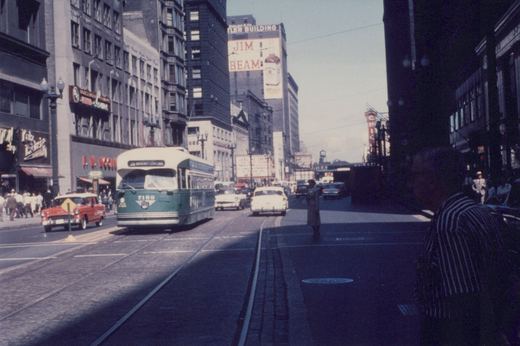 A green and cream streetcar approaches on double track in a busy downtown street lined with tall buildings, with 1950s cars, a Jim Beam wall sign and a movie marquee behind it.