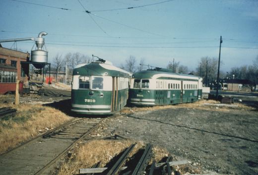 Two green and cream interurban cars, numbered 7210 and 7201, sit on tracks in a gravel yard with a clear sky and leafless trees in the background.