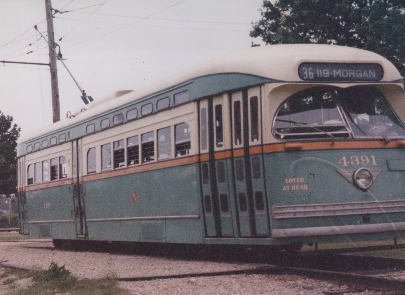 A green and cream streetcar with an orange stripe and a raised trolley pole stands on gravel track under overhead wire, its front sign reading 36 119-MORGAN.