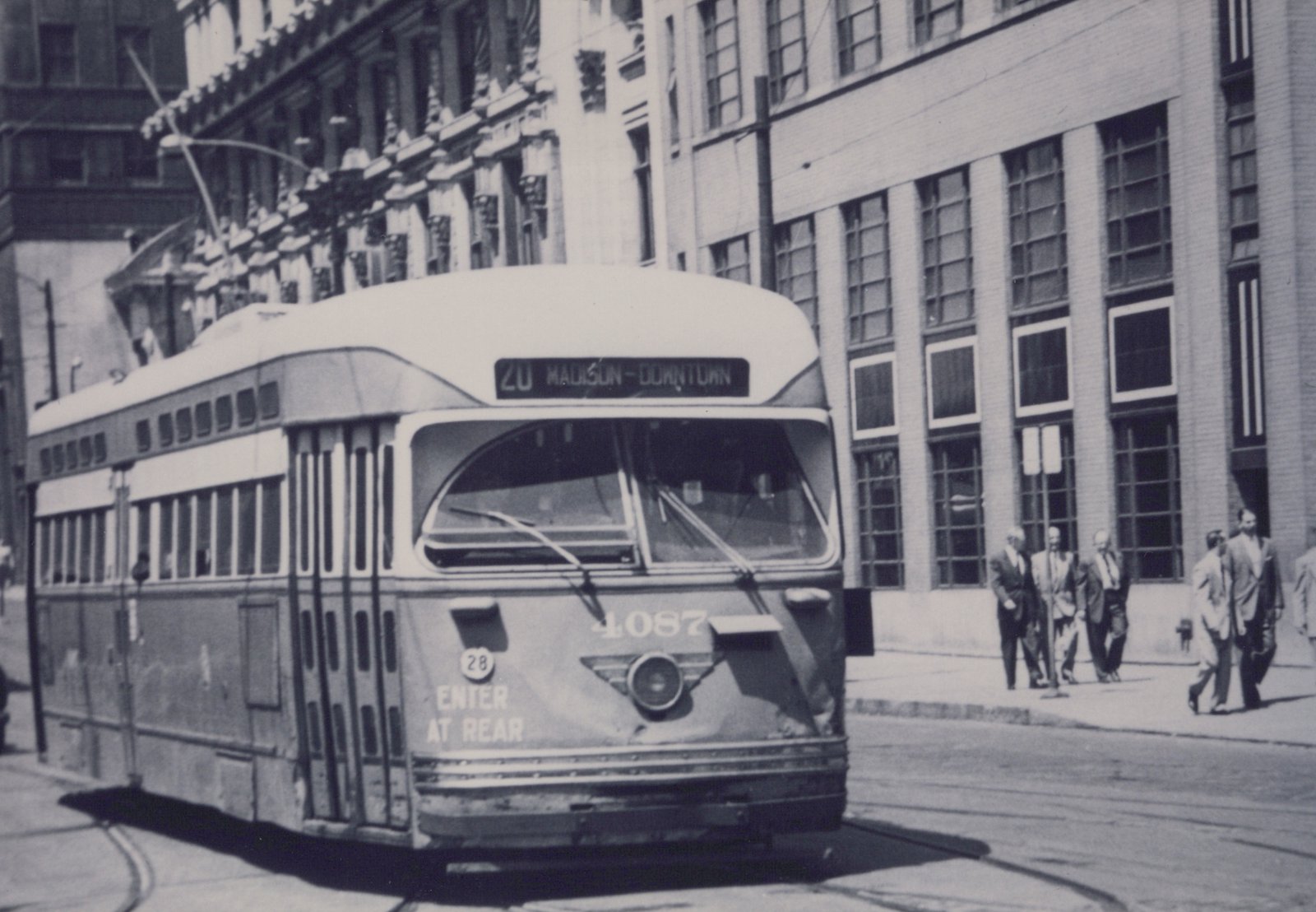 Black-and-white photo of a streamlined streetcar numbered 4087 with the sign 20 MADISON-DOWNTOWN, running on street tracks past office buildings while men in suits walk along the sidewalk.