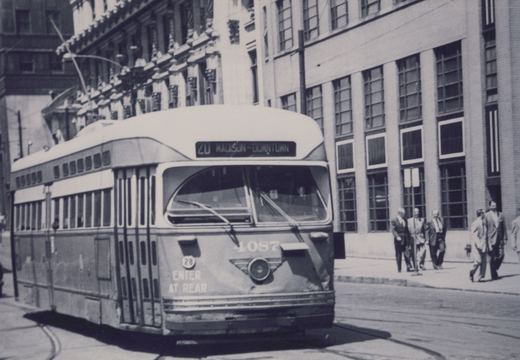 Black-and-white photo of a streamlined streetcar numbered 4087 with the sign 20 MADISON-DOWNTOWN, running on street tracks past office buildings while men in suits walk along the sidewalk.