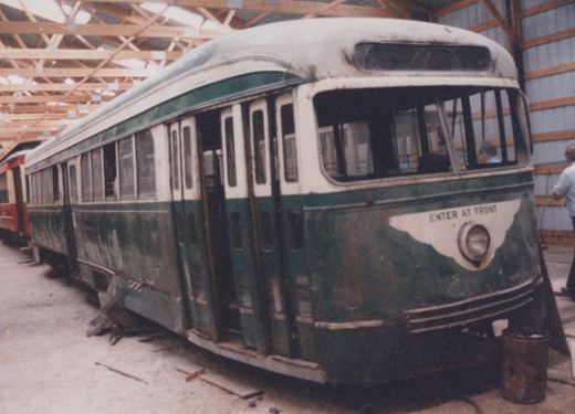 A rusted green and cream streamlined streetcar with broken windows and damaged lower side panels stands inside a wooden-trussed barn, with a red car behind it and a person standing at the right.