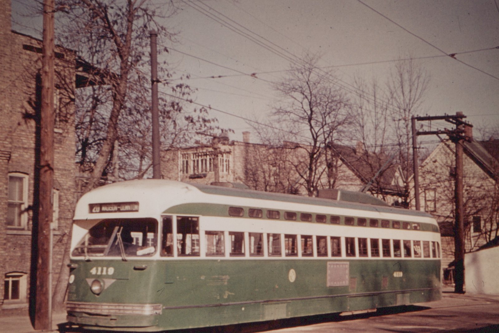 A green and cream streamlined streetcar with its trolley pole raised stands on a street beside utility poles, bare trees and brick buildings.