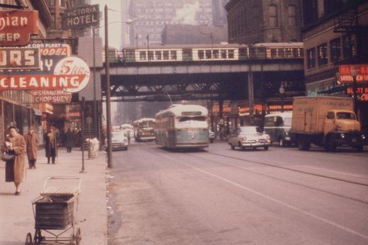 A green and cream streetcar comes toward the camera on a city street under a steel elevated structure carrying a two-car train, with neon signs, a yellow truck, a sedan and pedestrians in winter coats.