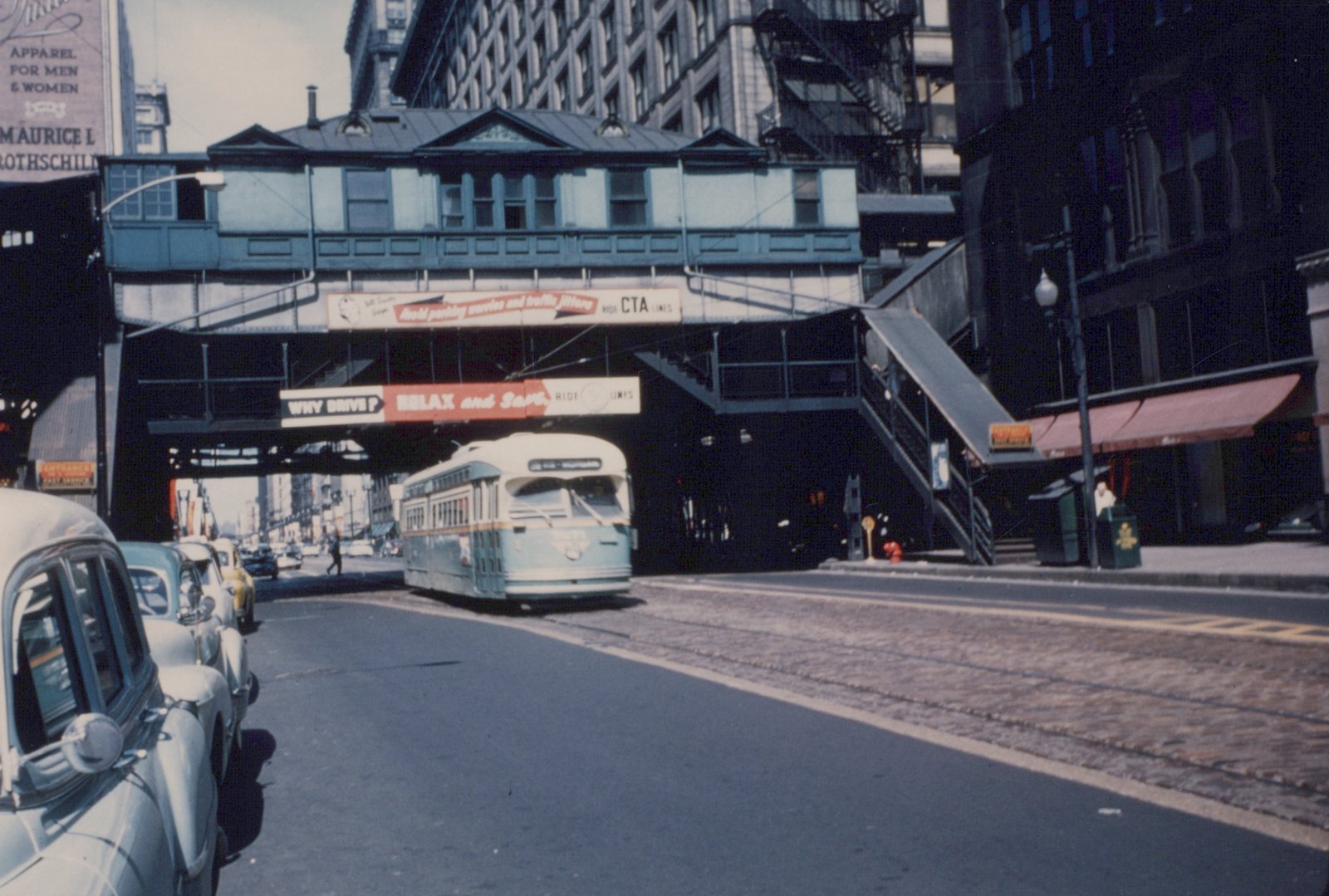 A light green and cream streetcar on cobblestone-paved tracks under an elevated railway station, with parked cars along the curb at left and tall downtown buildings behind.