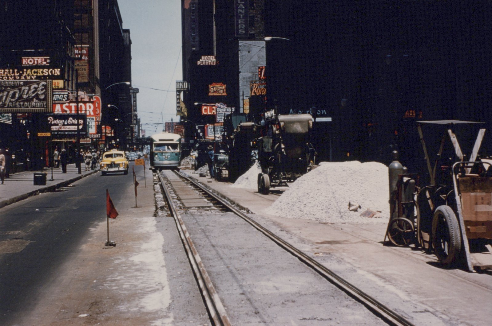 Looking down a downtown street lined with neon signs, a green and cream streetcar comes toward the camera on a single track that is under repair, with red warning flags, piles of crushed stone and construction machines beside it, and a yellow taxi in the next lane.