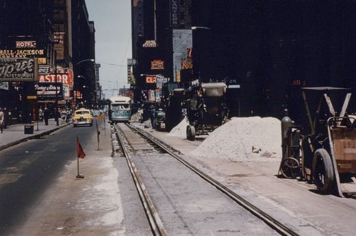 Looking down a downtown street lined with neon signs, a green and cream streetcar comes toward the camera on a single track that is under repair, with red warning flags, piles of crushed stone and construction machines beside it, and a yellow taxi in the next lane.