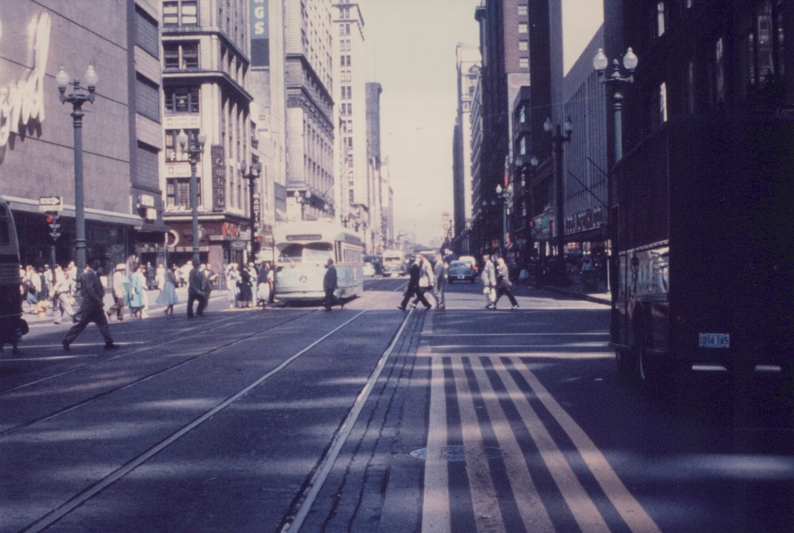 A light-colored streamlined streetcar stops at a busy downtown corner under tall buildings, with many pedestrians crossing the tracks in the foreground.