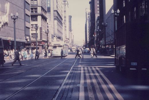 A light-colored streamlined streetcar stops at a busy downtown corner under tall buildings, with many pedestrians crossing the tracks in the foreground.