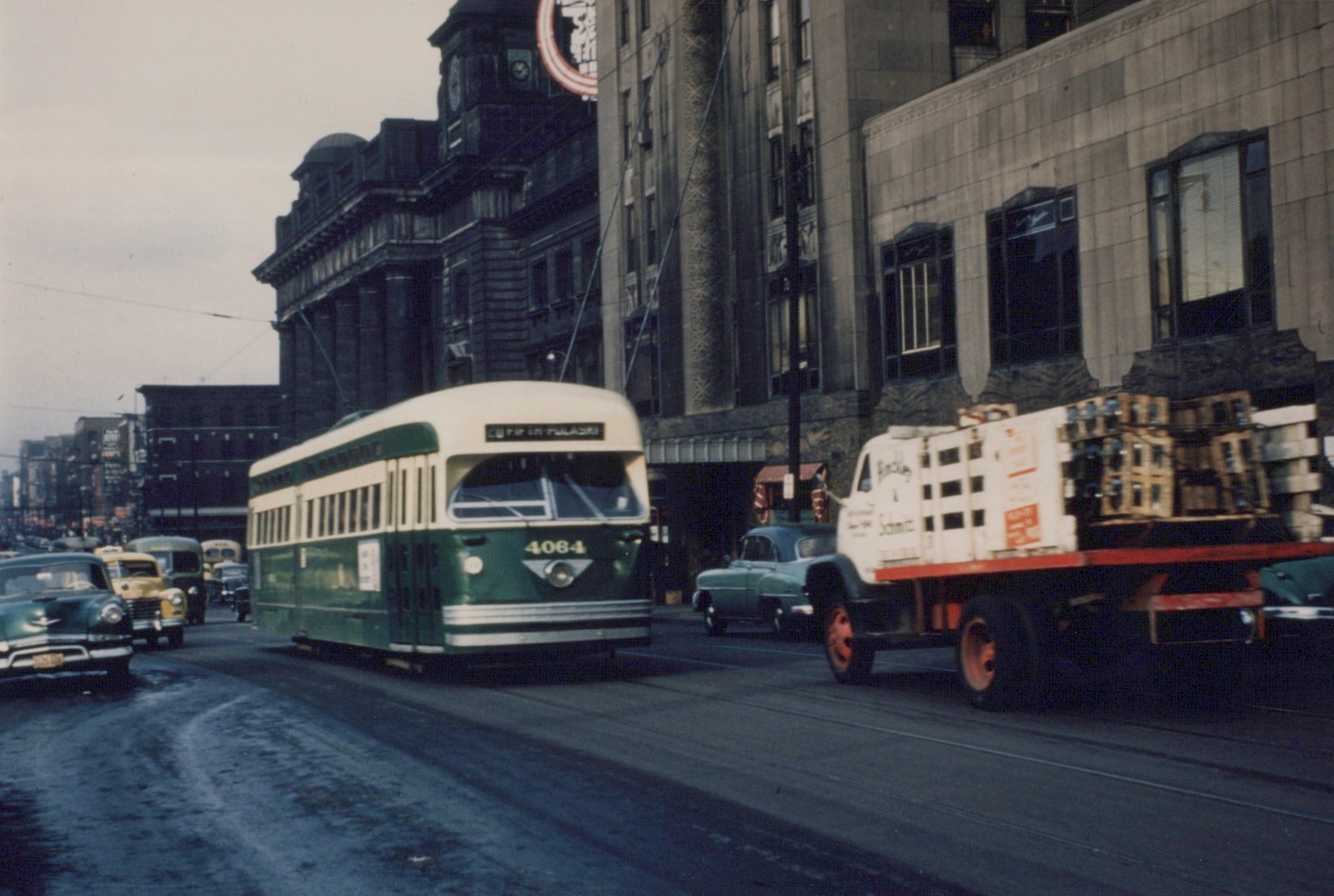 A green and cream streetcar numbered 4064 moves along a wet city street between early-1950s cars and a delivery truck loaded with bottle crates, with large stone buildings and a clock tower behind.