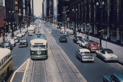A green and cream streetcar on center tracks in a downtown street lined with tall buildings, ornate lamp posts and 1950s cars, seen from above.
