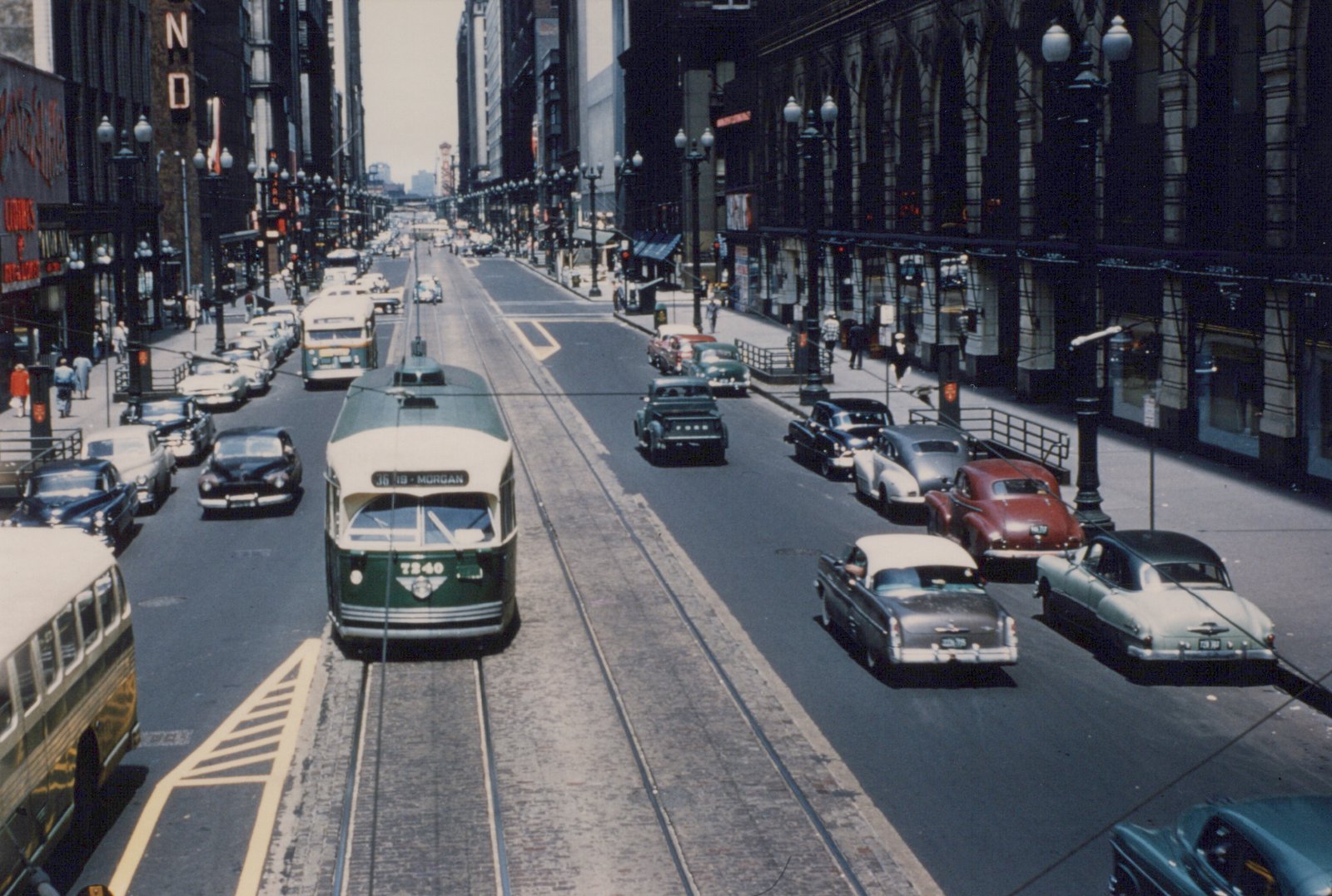 A green and cream streetcar on center tracks in a downtown street lined with tall buildings, ornate lamp posts and 1950s cars, seen from above.