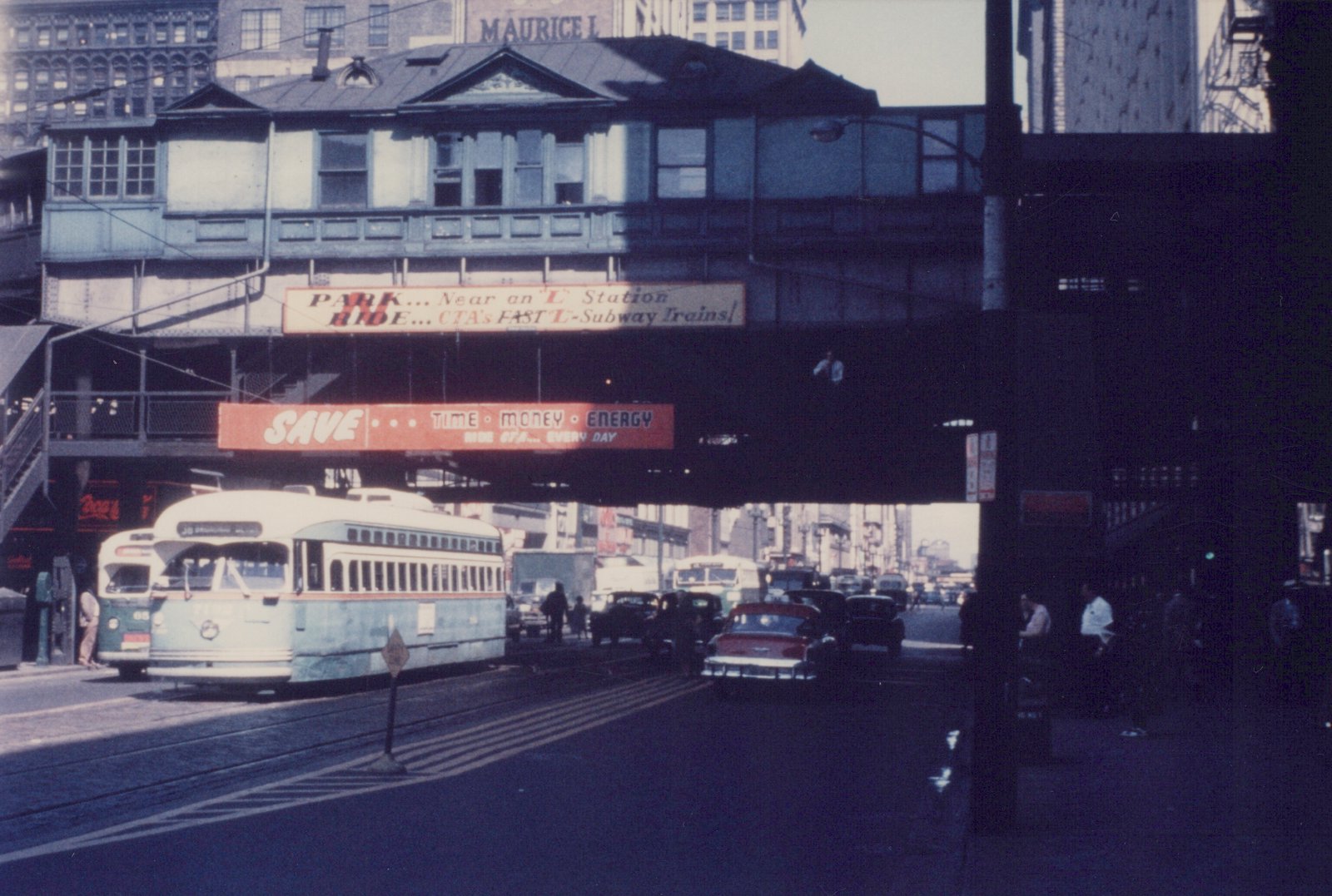 A green and cream streetcar sits under an elevated railway station that carries red and cream CTA banners, with a second streetcar beside it and traffic going off into the distance.