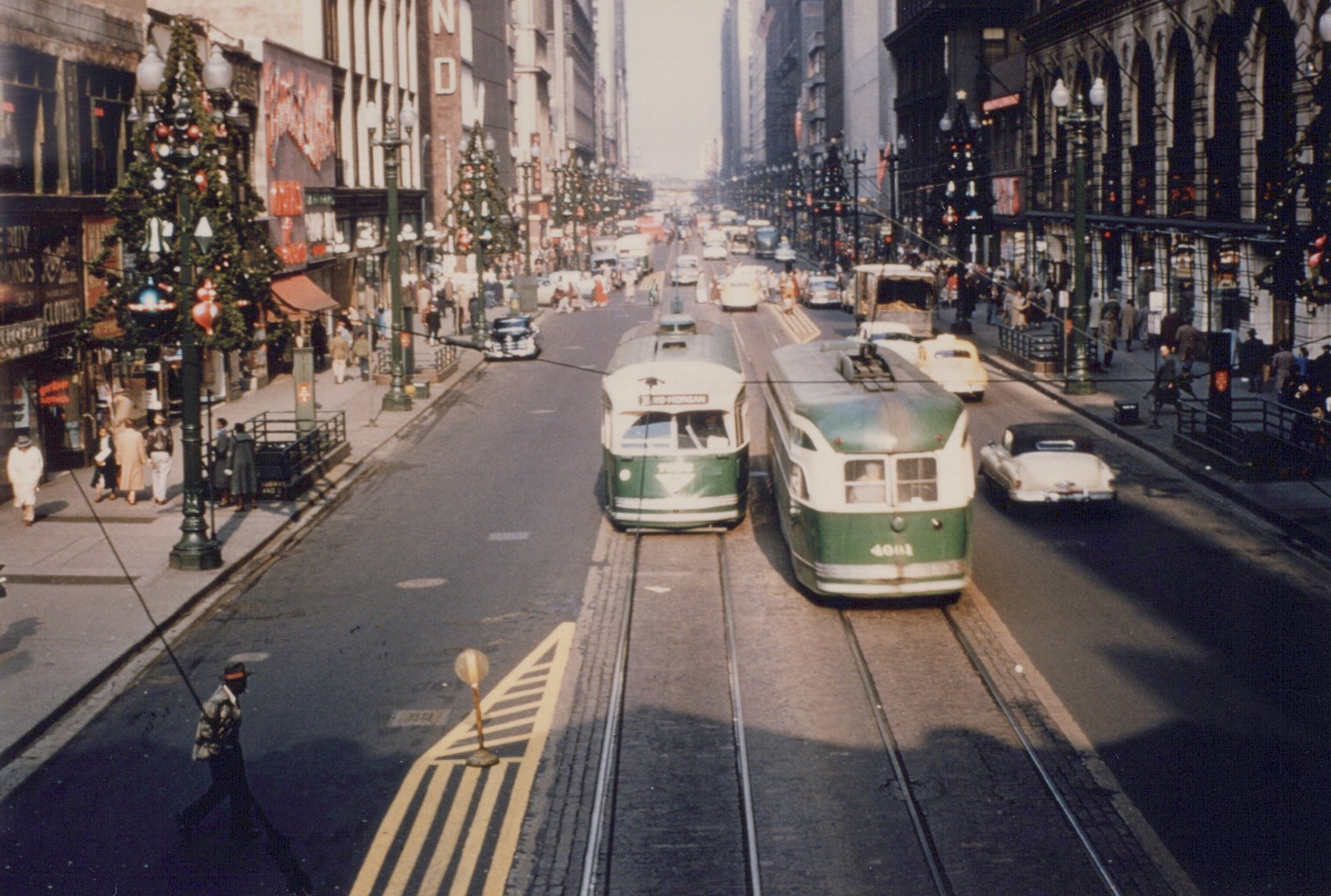 An elevated view down a busy downtown street decorated with Christmas trees on the lamp posts, with a green and cream streetcar coming toward the camera and another going away on the next track, among cars, shoppers and subway entrances.