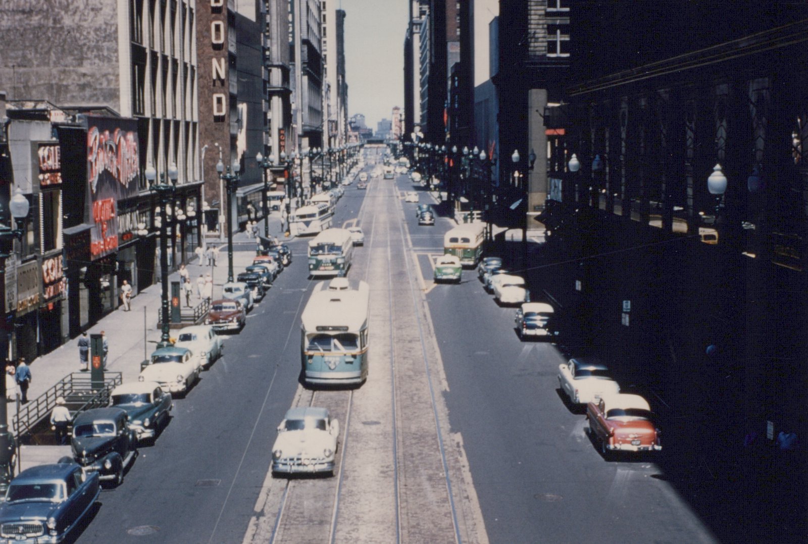 A high view down a downtown street lined with tall buildings and lamp posts. A green and cream streetcar runs on center tracks among parked cars, buses and pedestrians.