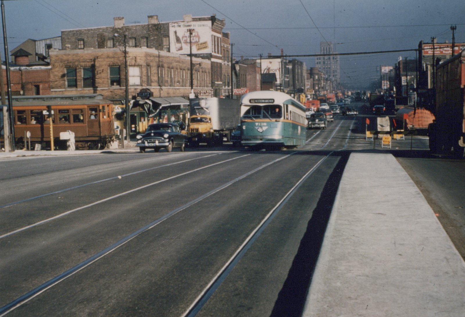 A green and cream streetcar comes toward the camera on a wide street with streetcar tracks; to the left, a brown wooden train car stands beside a brick tavern building, with a truck and a dark 1950s sedan between them.
