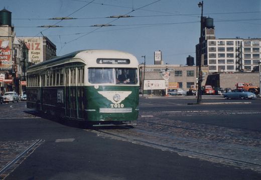 A green and cream streetcar numbered 7010, its sign reading 4 Cottage Grove, turns across a cobblestone street under overhead trolley wires, with brick buildings, billboards, a gas station and parked 1950s cars and trucks behind it.