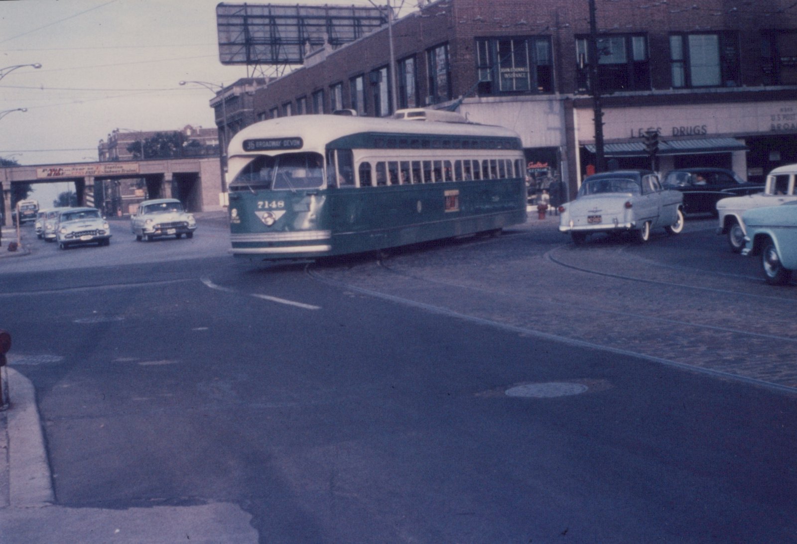 A green and cream streetcar signed 36 Broadway-Devon turns across a brick-paved intersection, with 1950s cars, a corner drug store and an overpass behind it.
