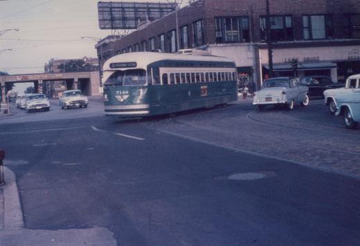 A green and cream streetcar signed 36 Broadway-Devon turns across a brick-paved intersection, with 1950s cars, a corner drug store and an overpass behind it.
