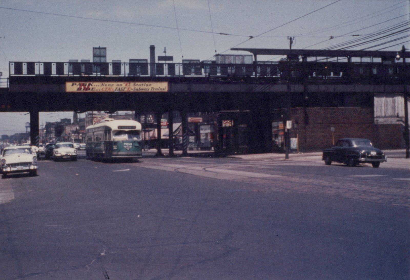 A green and cream streetcar comes toward the camera on a wide street, under a steel elevated railway bridge that carries a CTA 'Park and Ride' sign, with 1950s cars on both sides.