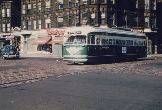 A green and cream streetcar with a trolley pole crosses a cobblestone intersection with tracks in front of a brick corner building with a Walgreens store, and a dark sedan is parked at left.
