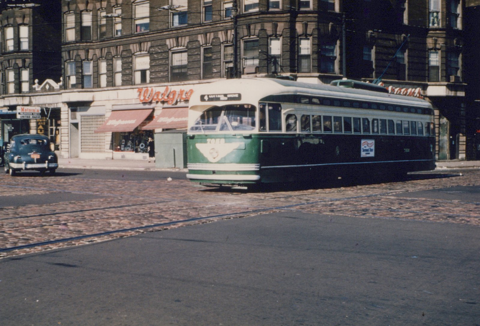 A green and cream streetcar with a trolley pole crosses a cobblestone intersection with tracks in front of a brick corner building with a Walgreens store, and a dark sedan is parked at left.