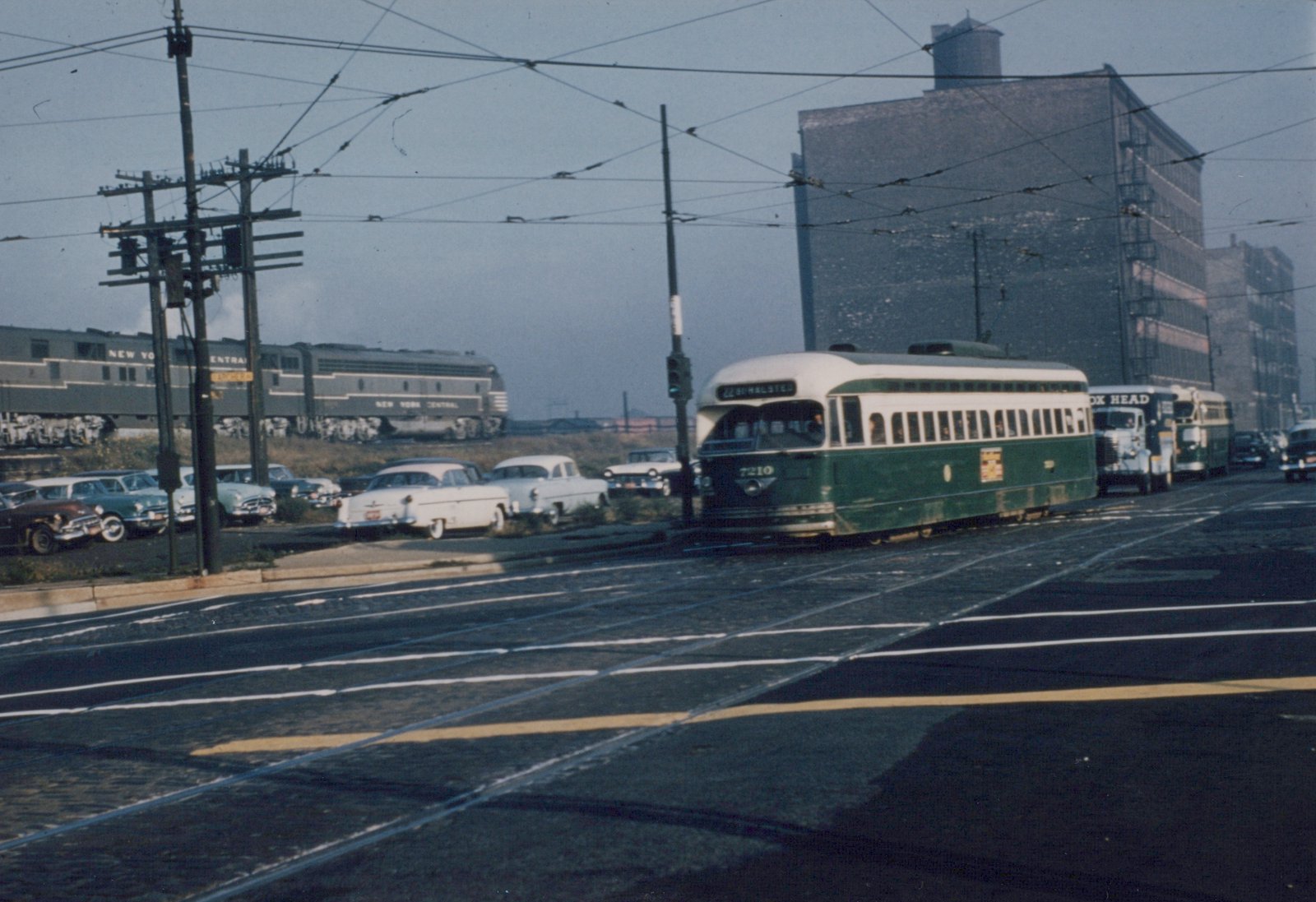 A green and cream streetcar on a cobbled street under trolley wires. A truck and a bus follow it, a gray diesel passenger train runs on an embankment behind parked 1950s cars, and brick warehouses stand at right.