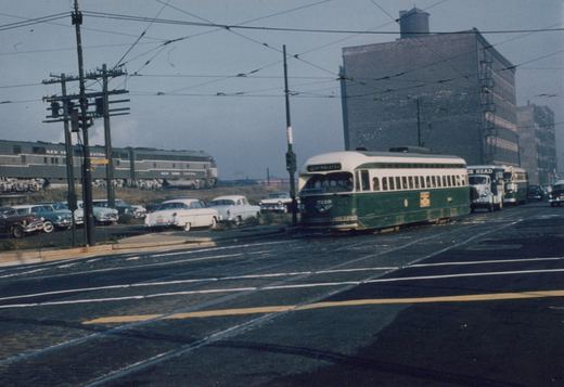 A green and cream streetcar on a cobbled street under trolley wires. A truck and a bus follow it, a gray diesel passenger train runs on an embankment behind parked 1950s cars, and brick warehouses stand at right.
