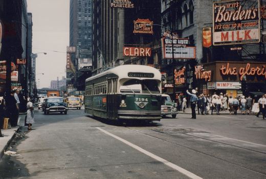 A green and cream streetcar signed 22 81-HALSTED comes up a downtown street under bright neon signs for Borden's Milk, the Blue Note and Wimpy, while a traffic officer directs a crowd of pedestrians.