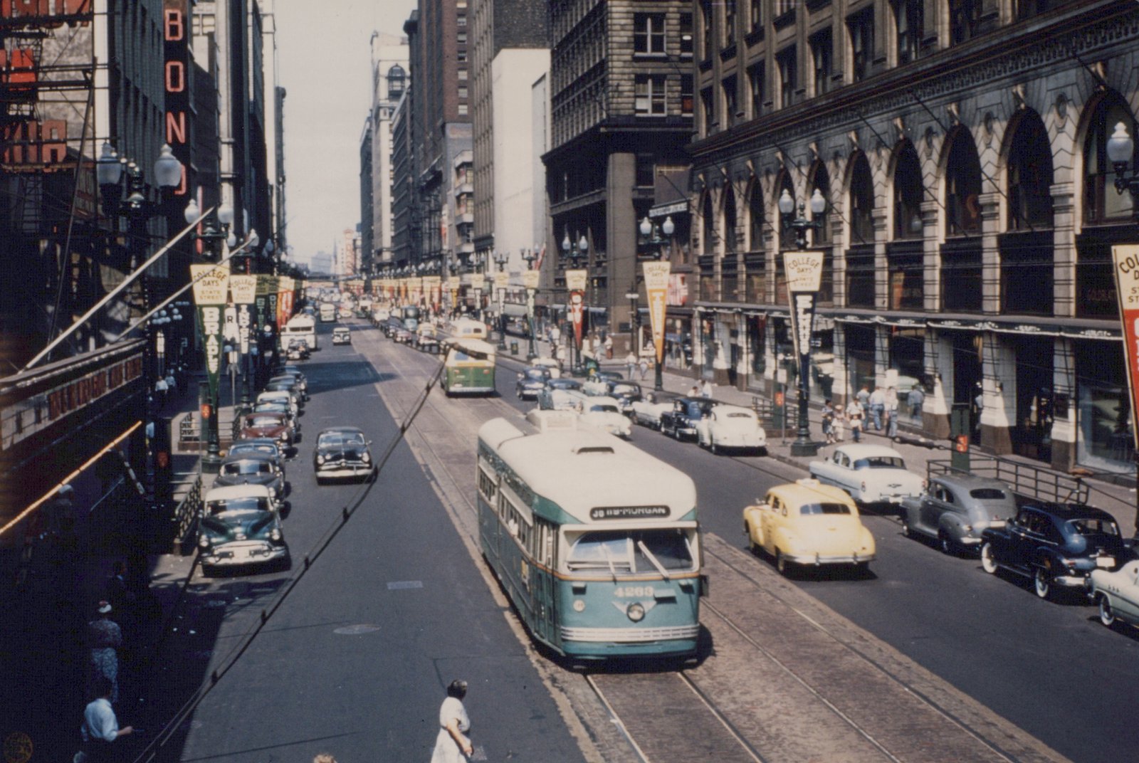 A green and cream streetcar on tracks in the middle of a busy downtown street lined with tall buildings, parked cars, a yellow taxi and pennant banners on the lamp posts.
