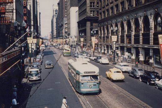 A green and cream streetcar on tracks in the middle of a busy downtown street lined with tall buildings, parked cars, a yellow taxi and pennant banners on the lamp posts.