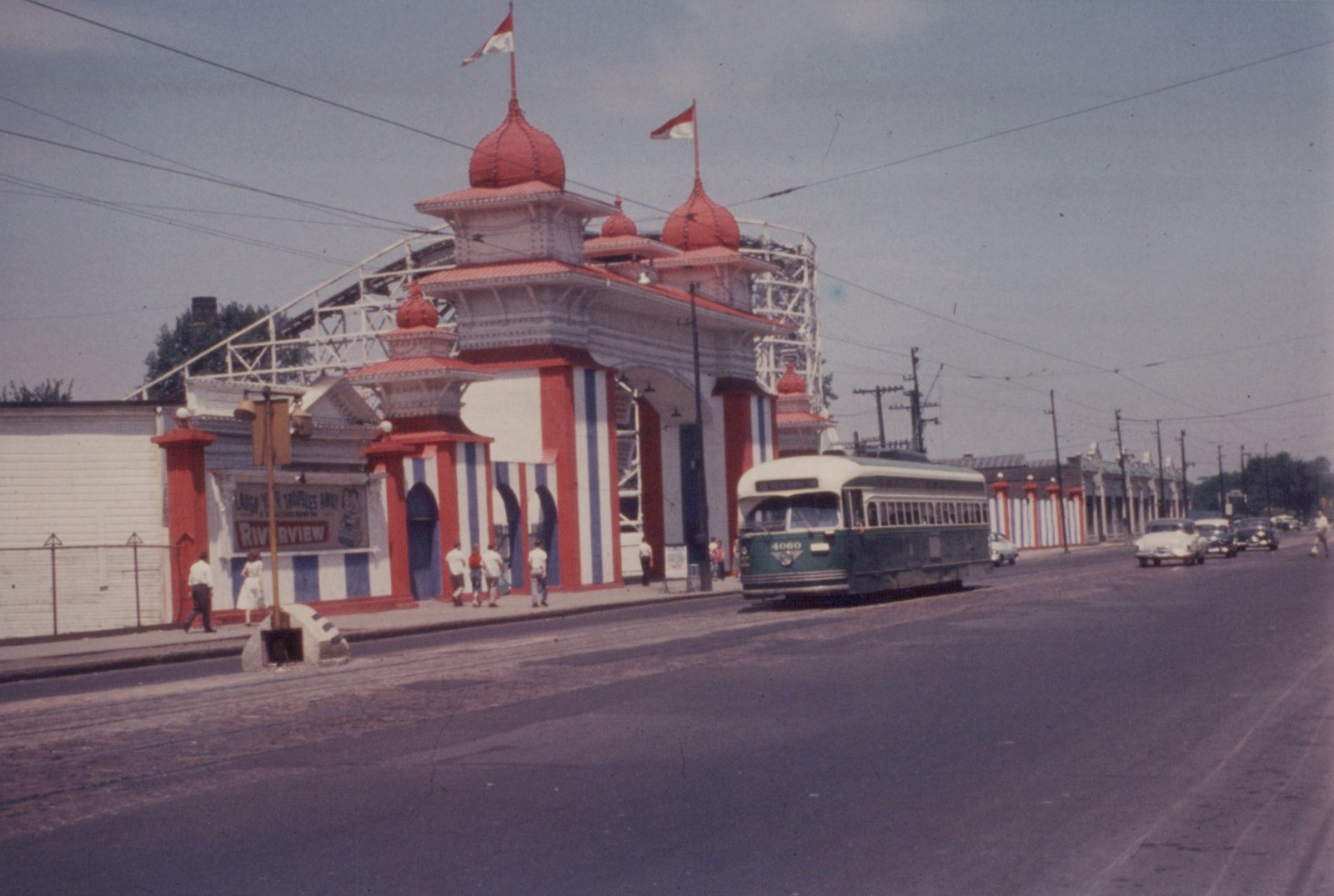 A green and cream streetcar on a wide street passes an ornate red, white and blue amusement park gate with onion domes, flags and a roller coaster behind it, while pedestrians walk along the sidewalk.