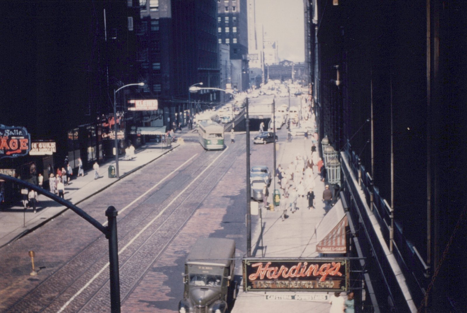 Elevated view down a busy downtown Chicago street with a green and cream streetcar on double track, crowded sidewalks, a mail truck in the foreground and a neon Harding's sign at lower right.