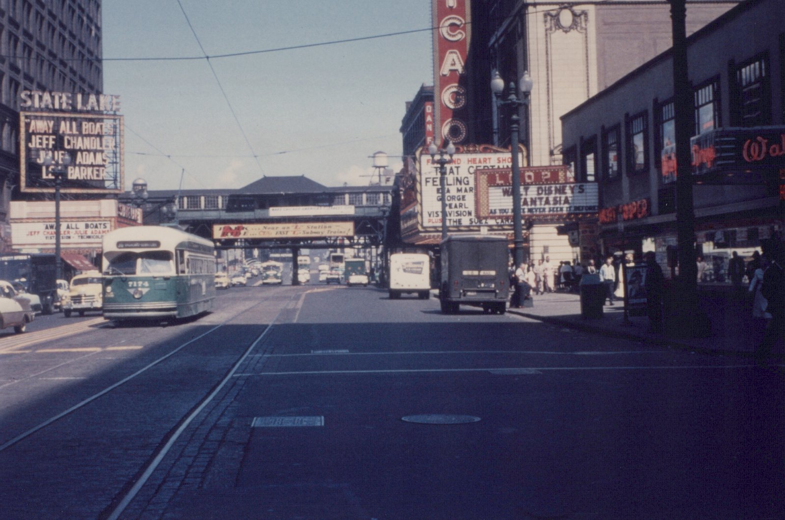 A green and cream streetcar on a wide downtown street lined with theater marquees, with an elevated railway crossing the street in the distance.