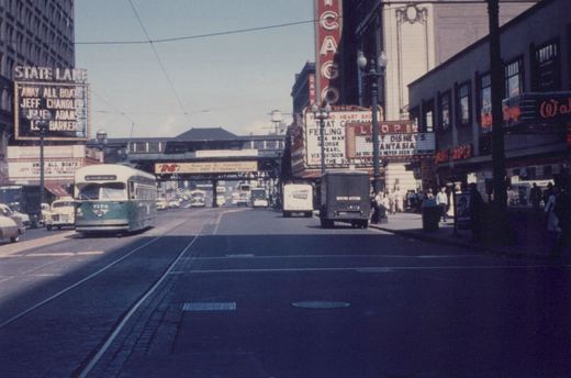 A green and cream streetcar on a wide downtown street lined with theater marquees, with an elevated railway crossing the street in the distance.